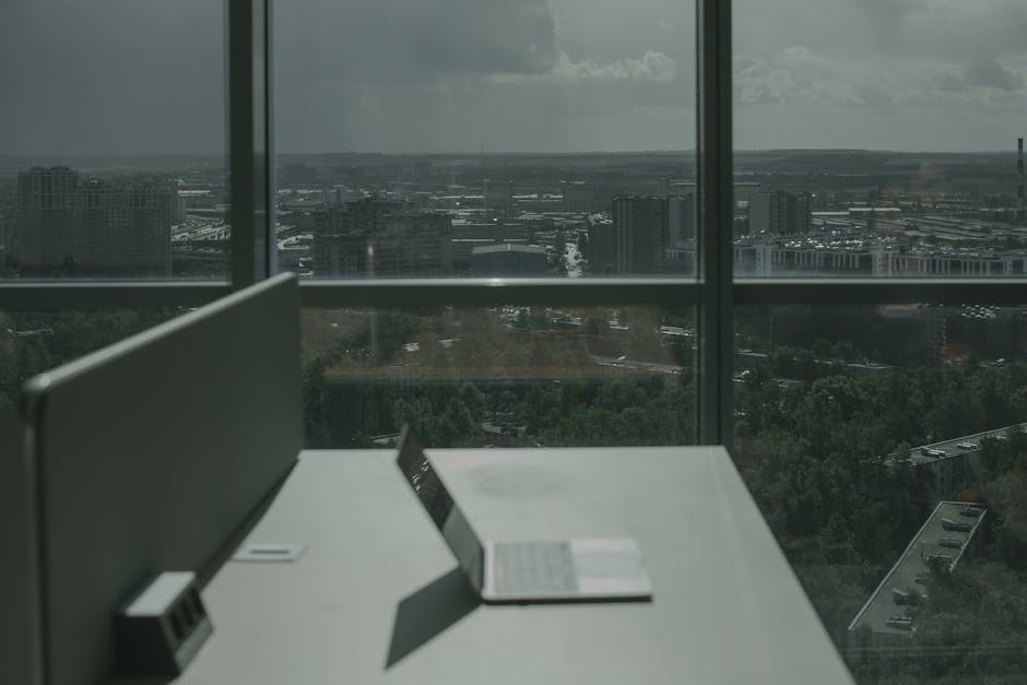 Laptop on office desk with a city skyline through a large window, offering a modern workspace view.